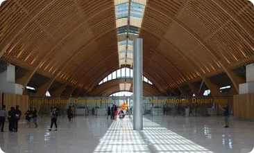 写真：大きな木造アーチの天井が特徴的な、国際線出発ゲートの広々とした空港ロビー。自然光が差し込み、明るく洗練された空間の中を歩く人々の姿が見られる。