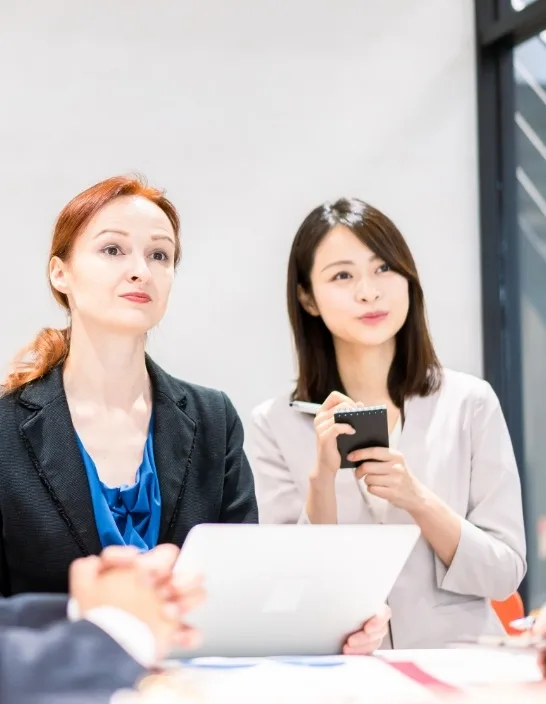 写真：2人の女性が会議室でノートパソコンを使いながら会話している。